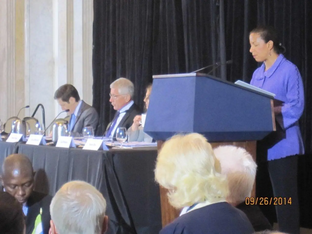 It is a conference, there is a woman standing in front of a table and speaking something, beside...