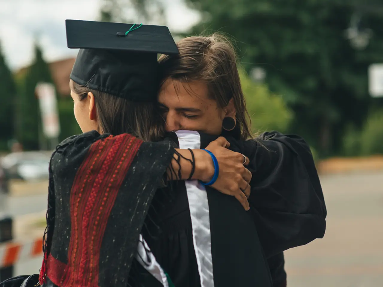 In the image there are two persons hugging each other in graduation costume and in the background...