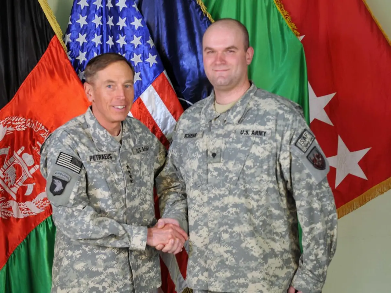 In this image there are two persons standing with a smile and they are shaken hands, behind them...