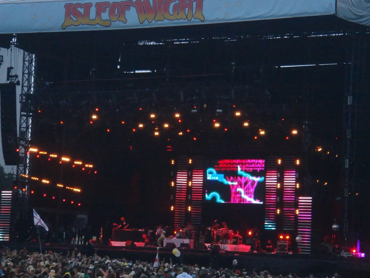 In this image we can see stage with lights. Also there is a screen. And there are people playing...