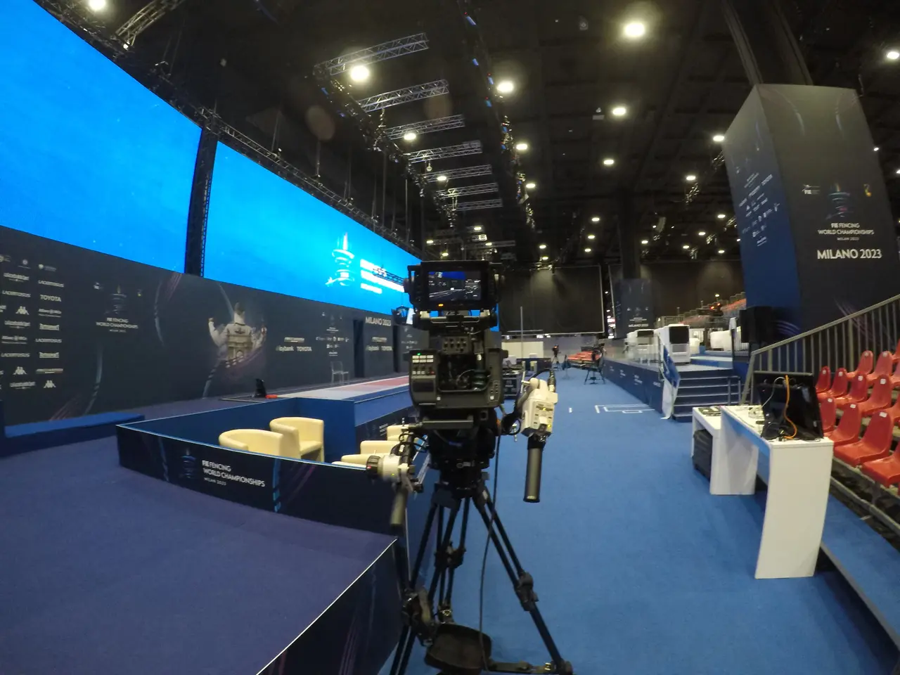 The image shows a video camera on a tripod in front of a large screen, surrounded by chairs,...