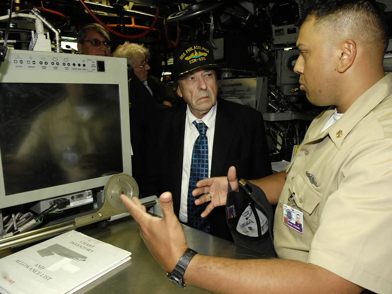 The image shows two men in military uniforms talking to each other in front of a computer, with a...
