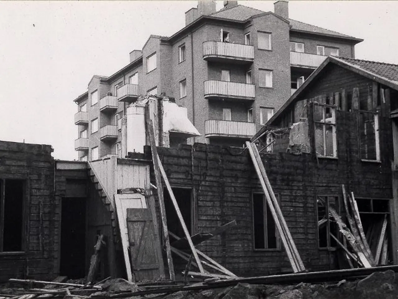 The image shows a black and white photo of a building that has been demolished, with wooden sticks...