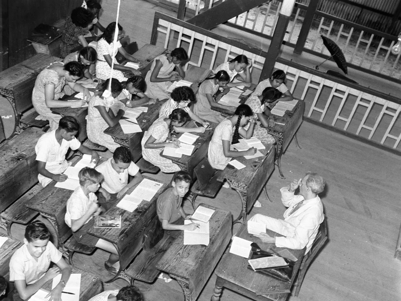 The image shows a black and white photo of a classroom full of children sitting at desks, with...