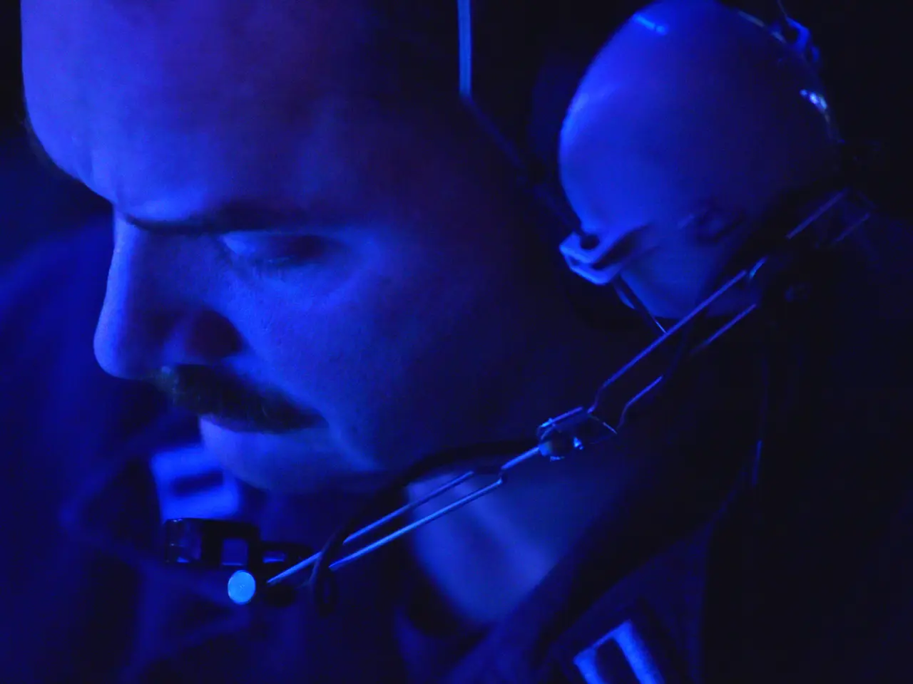 The image shows a man wearing headphones and a microphone in a dark room. He has a serious...