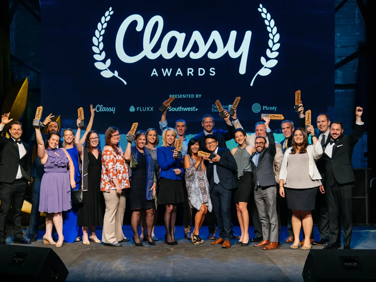 The image shows a group of people standing on top of a stage, smiling and holding awards in their...