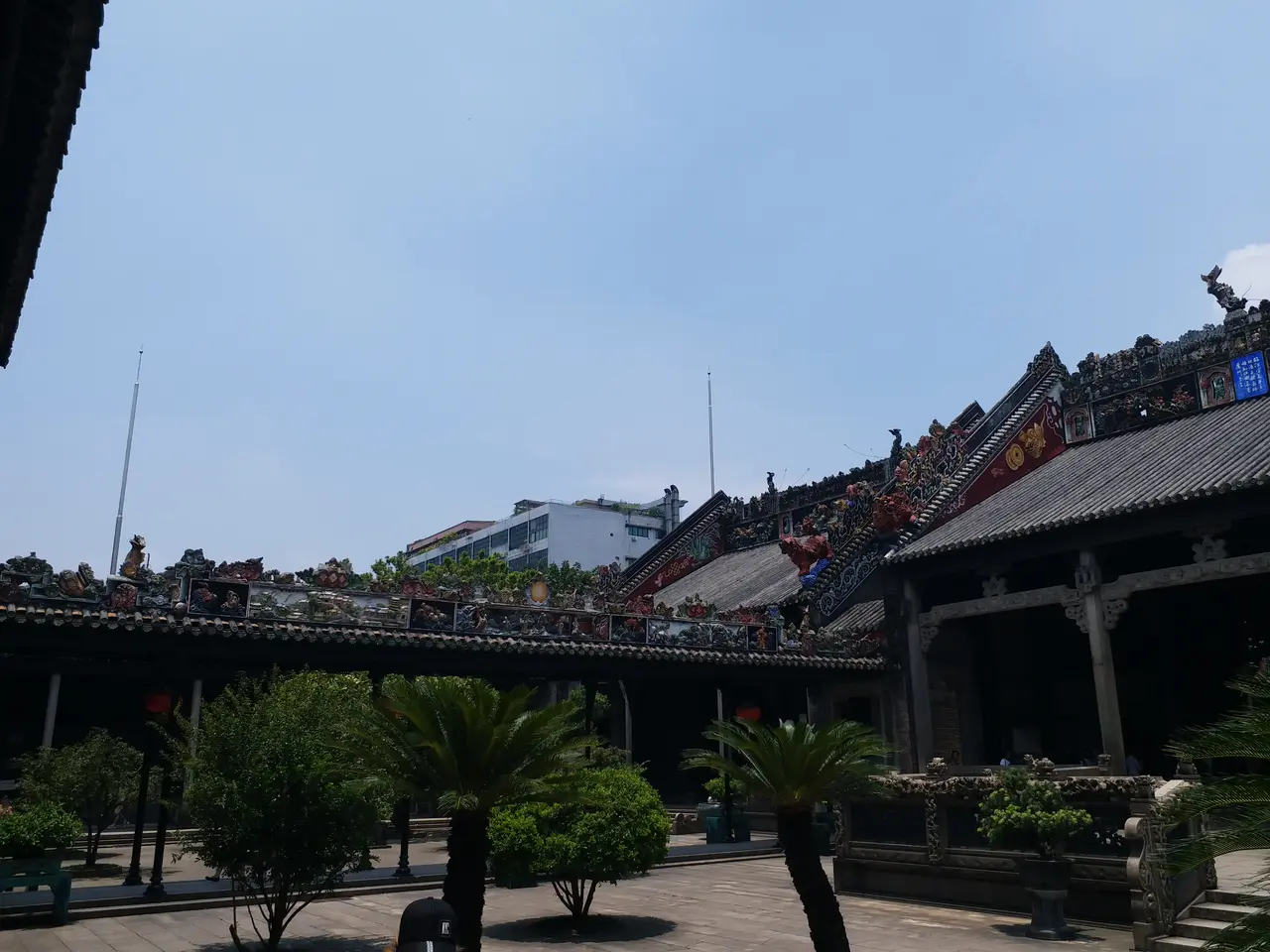 The image shows a large building with a lot of statues on top of it, surrounded by trees, plants,...