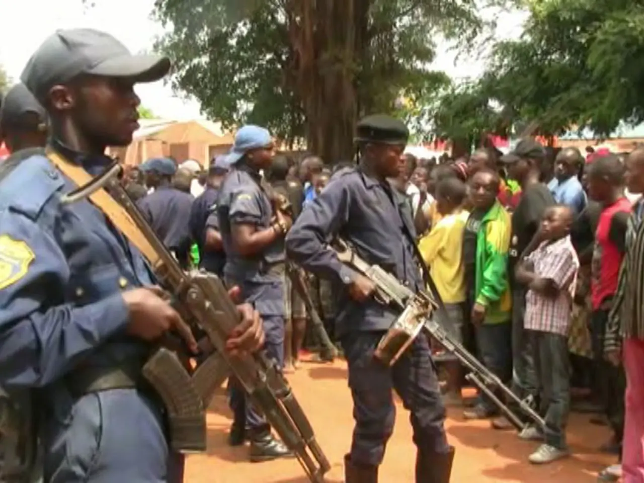 The image shows a group of police officers standing in front of a crowd of people, some of whom are...