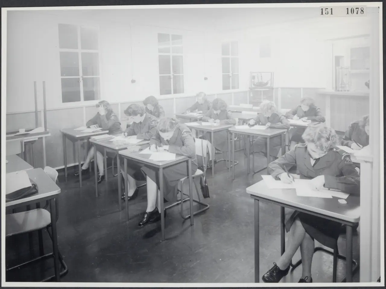 The image shows a black and white photo of a classroom full of students sitting at desks, writing...