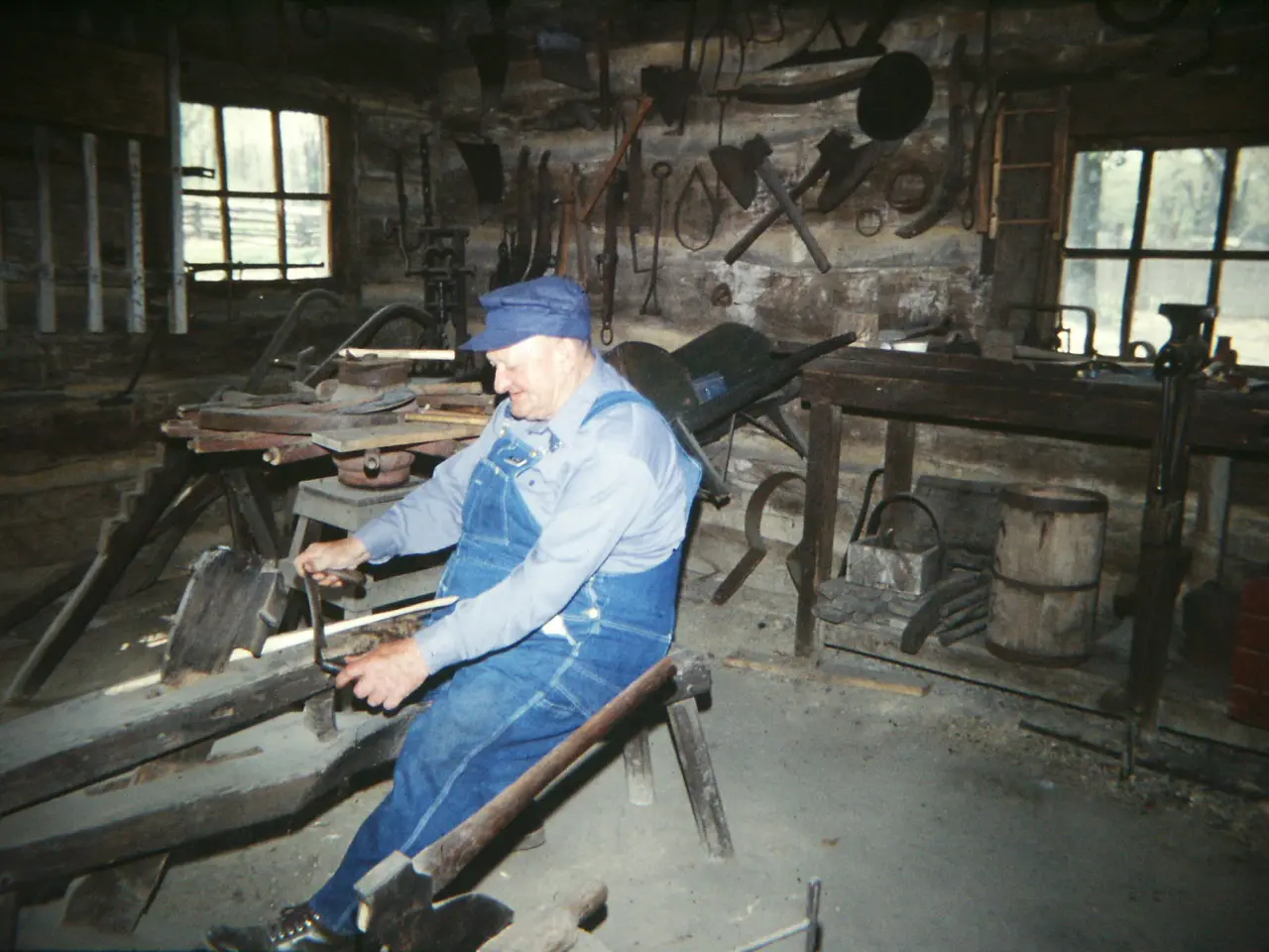 The image shows a blacksmith wearing a blue dress and cap, sitting on a bench and working on a...