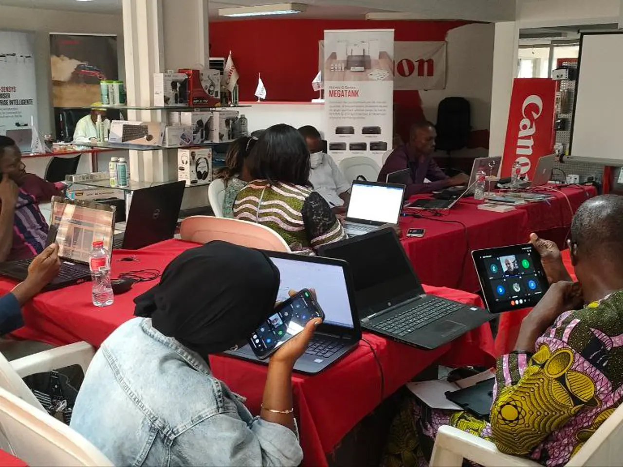 The image shows a group of people sitting around a table with laptops, bottles, and other objects....