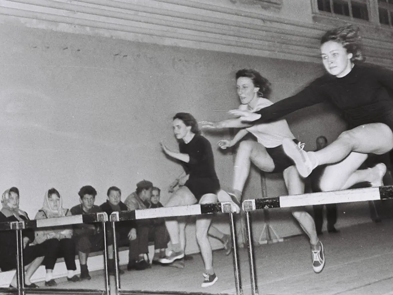 The image shows a black and white photo of two women jumping over a hurdle in a gymnasium. There...