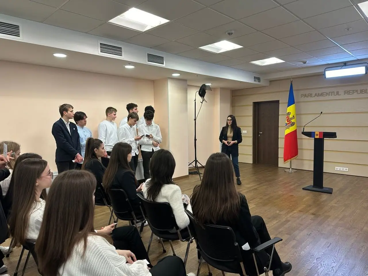 The image shows a group of people sitting in chairs in front of a podium, with a woman standing...