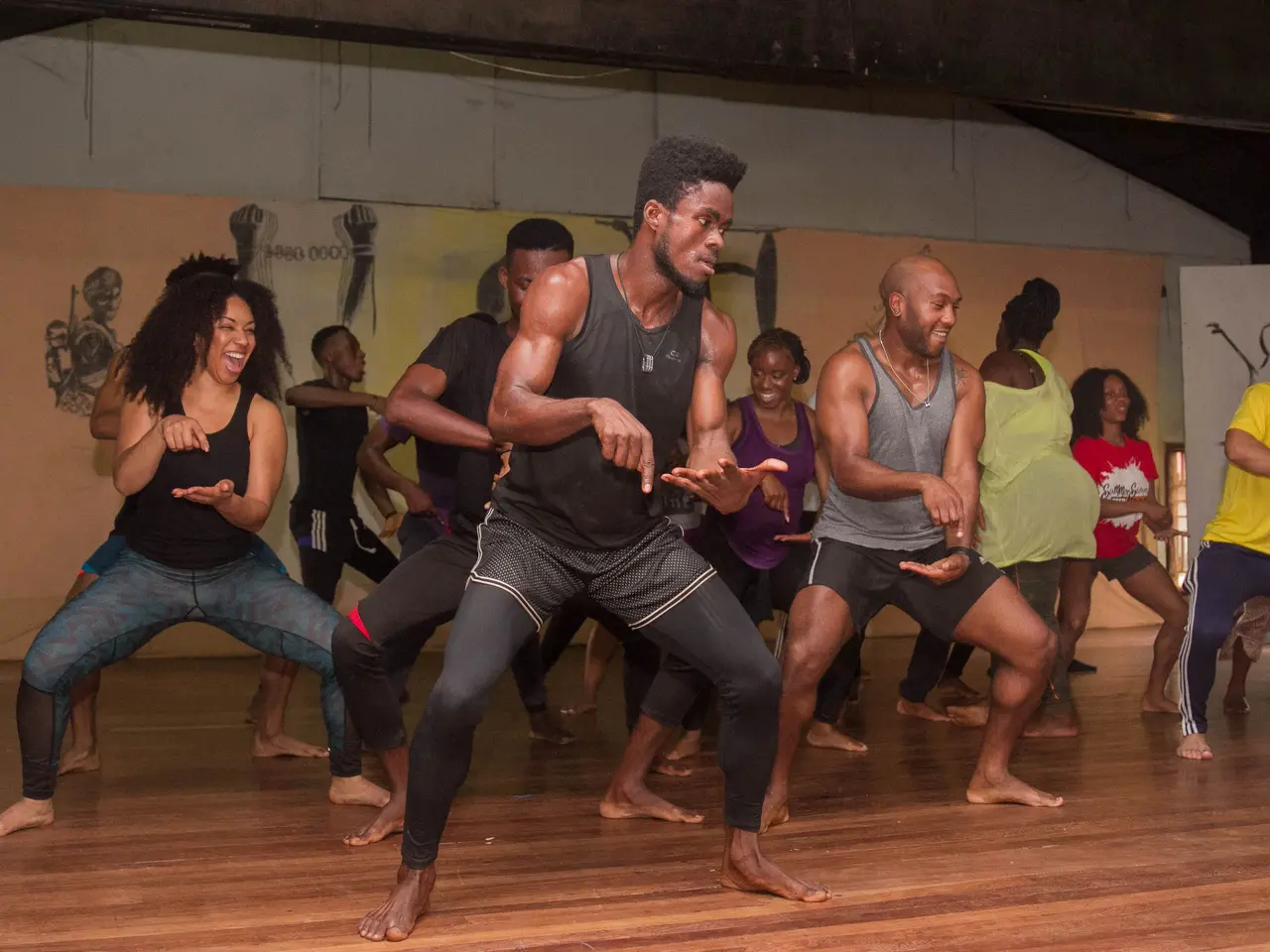 The image shows a group of people in a dance class in a room with a wall adorned with paintings in...
