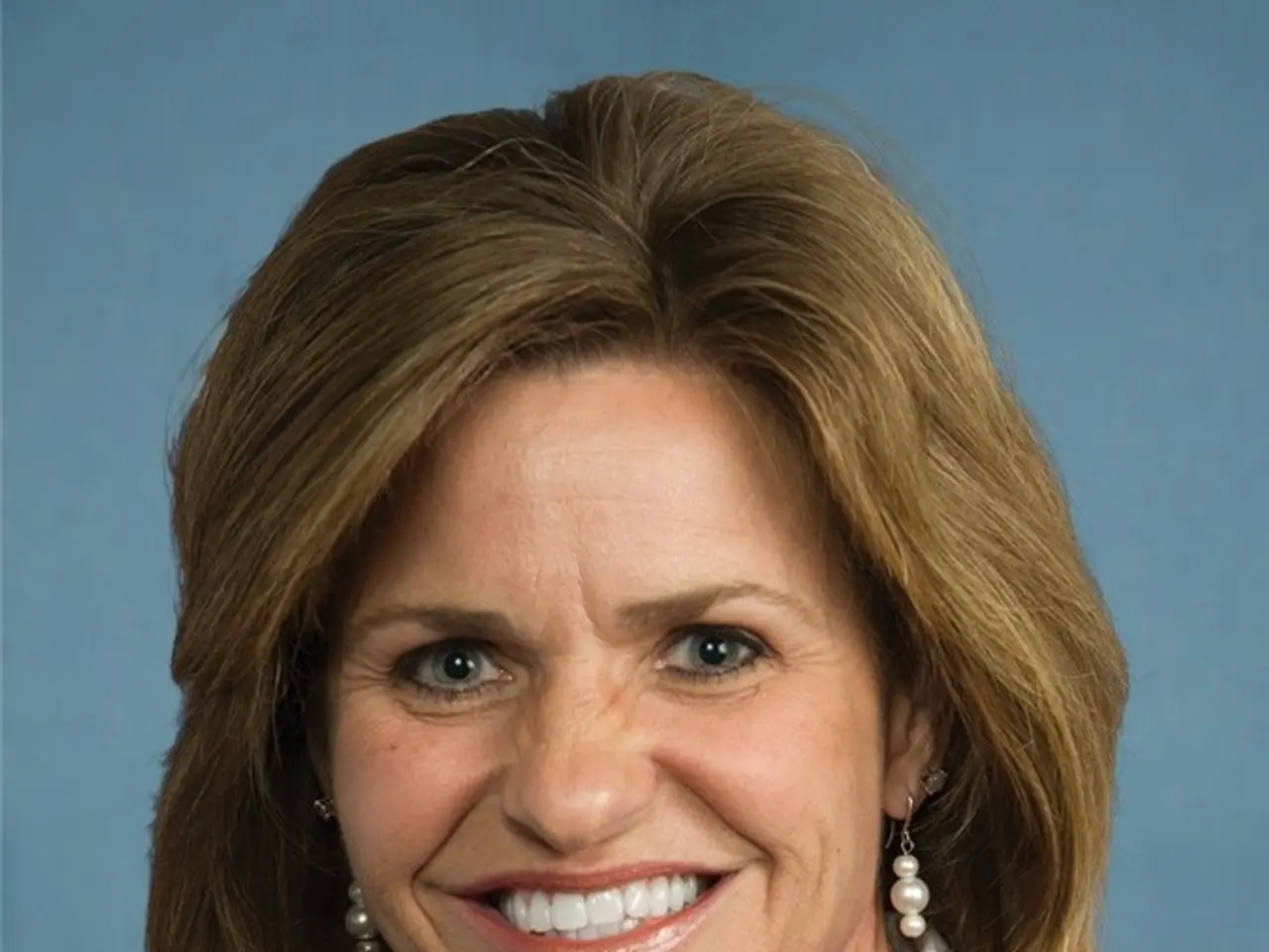 The image shows a woman in a business suit smiling at the camera against a blue background. She...