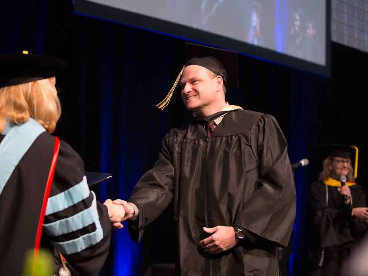 The image shows a man in a graduation cap and gown shaking hands with a woman in front of a podium,...
