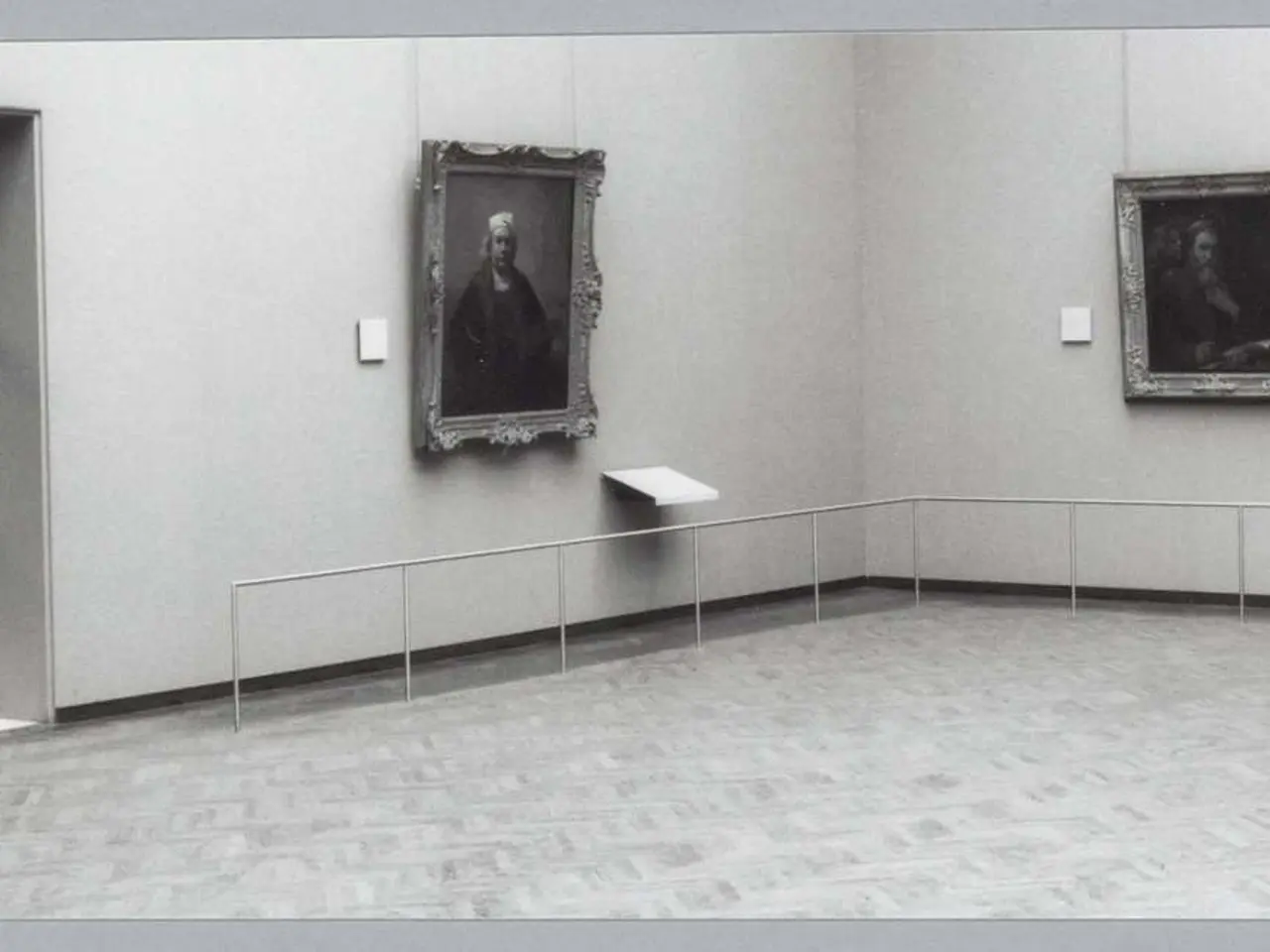 The image shows a black and white photo of a room with paintings on the walls, a floor, railings,...