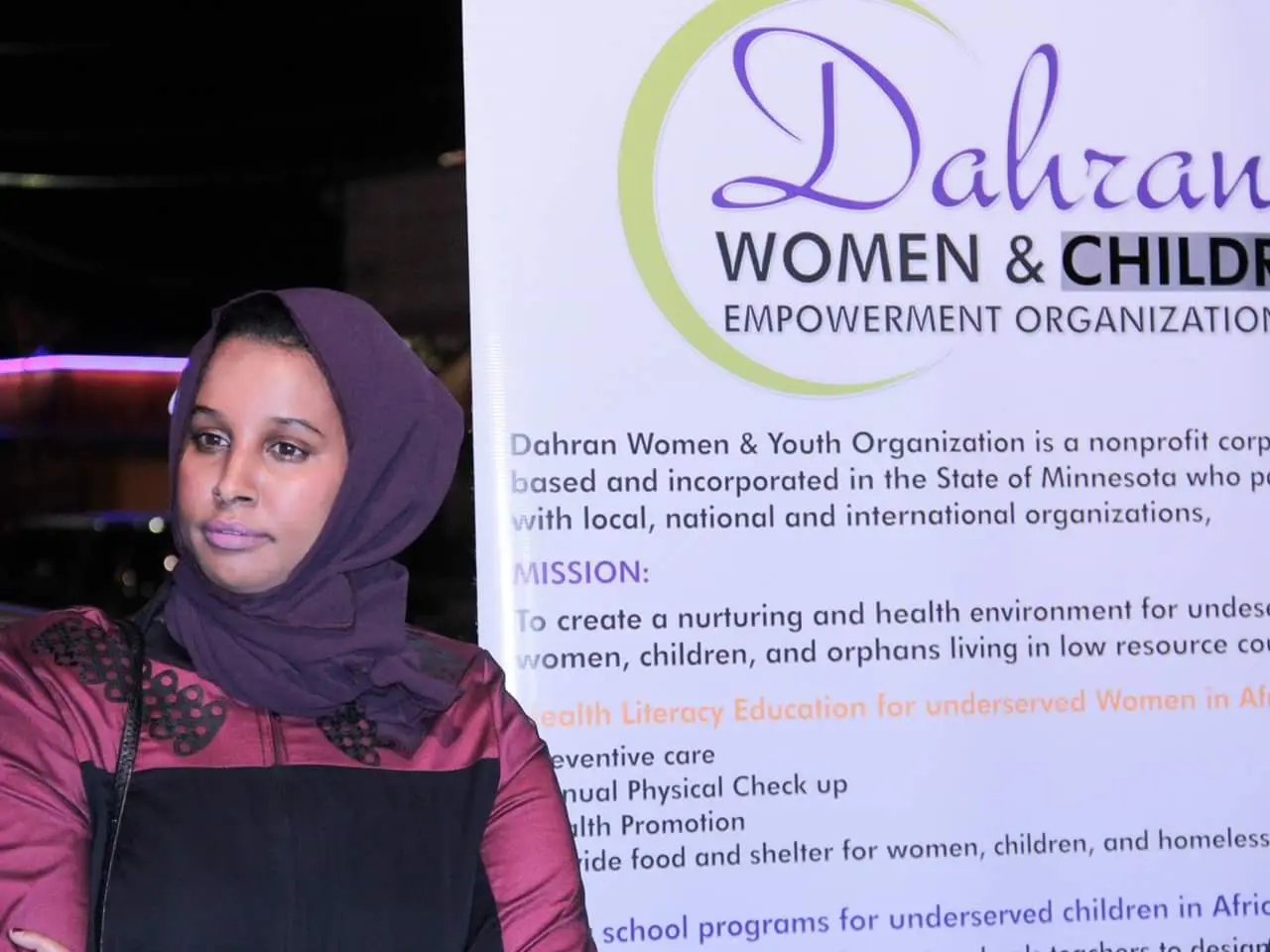 The image shows a woman standing in front of a banner that reads "Dahran Women & Children...