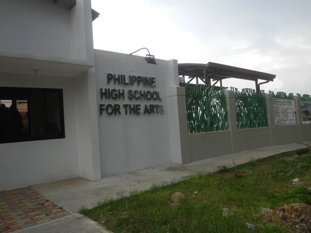 The image shows a building with windows, a wall with text on it, a roof with metal poles, a group...