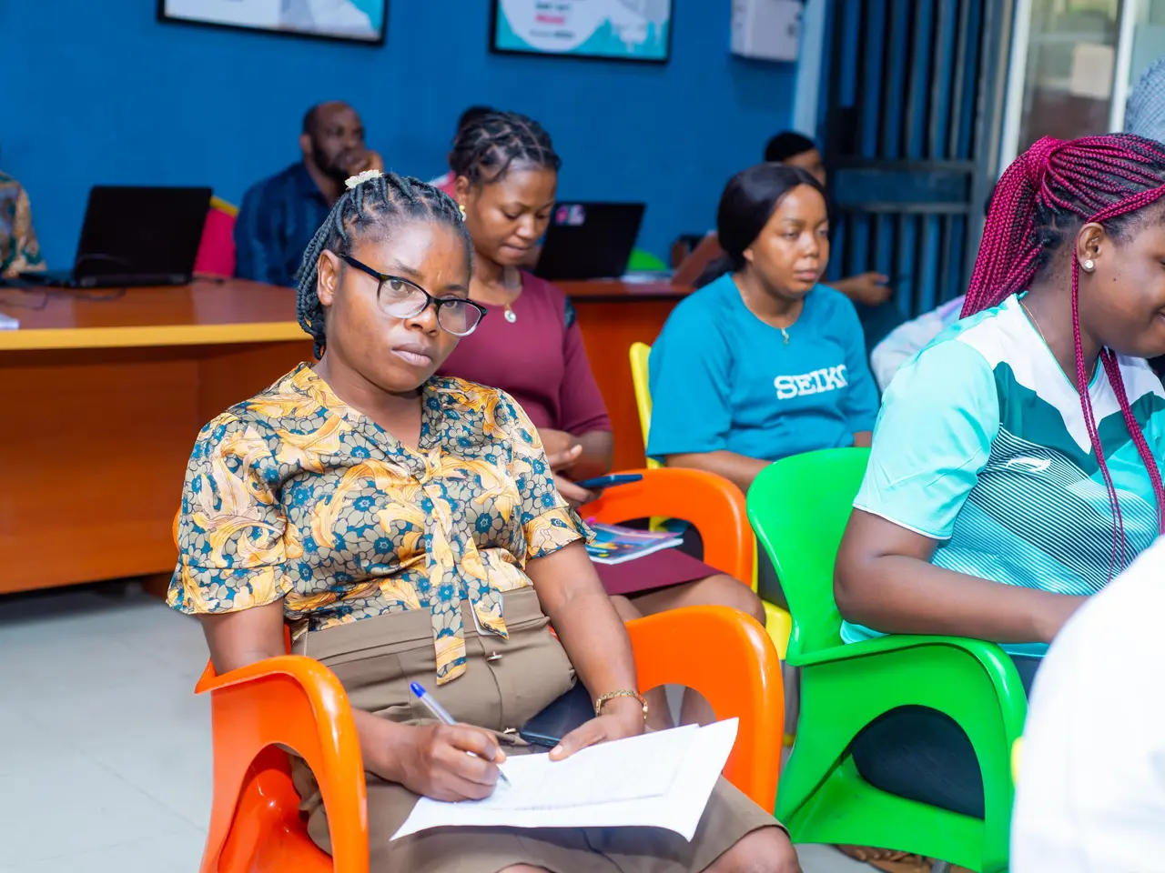 The image shows a group of people sitting in chairs in a room with laptops, books, and other...
