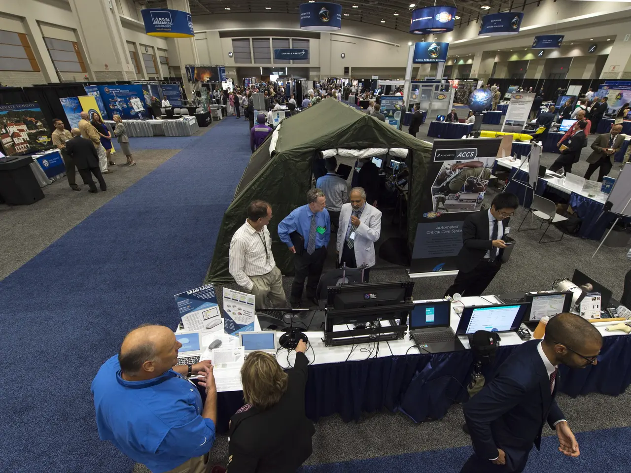The image shows a large group of people standing around a table with laptops, monitors, papers, and...