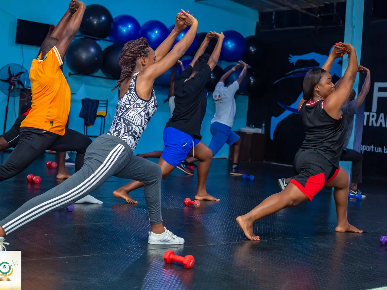 The image shows a group of people doing yoga in a gym, with dumbbells scattered on the floor. In...