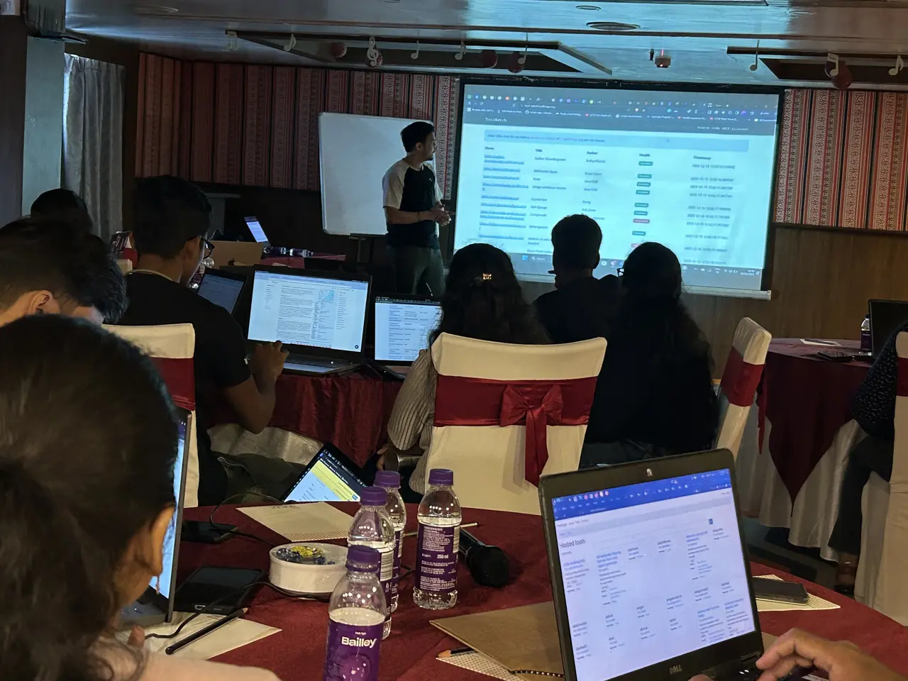 The image shows a group of people sitting around a table with laptops, bottles, papers, pens, and...
