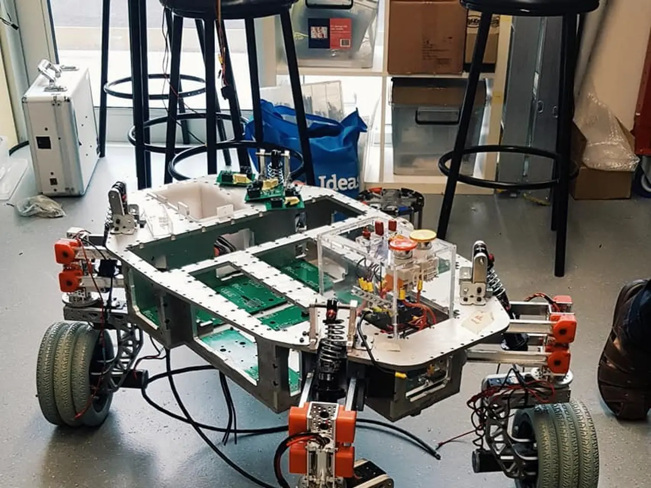 The image shows a robot sitting on top of a table in a room, surrounded by stools, cardboard boxes,...