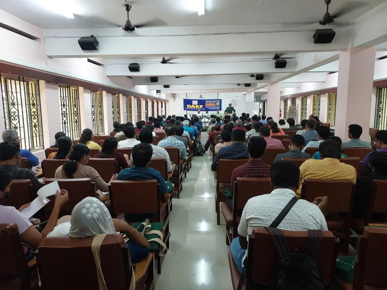 The image shows a large group of people sitting in chairs in a room with windows on the left and...