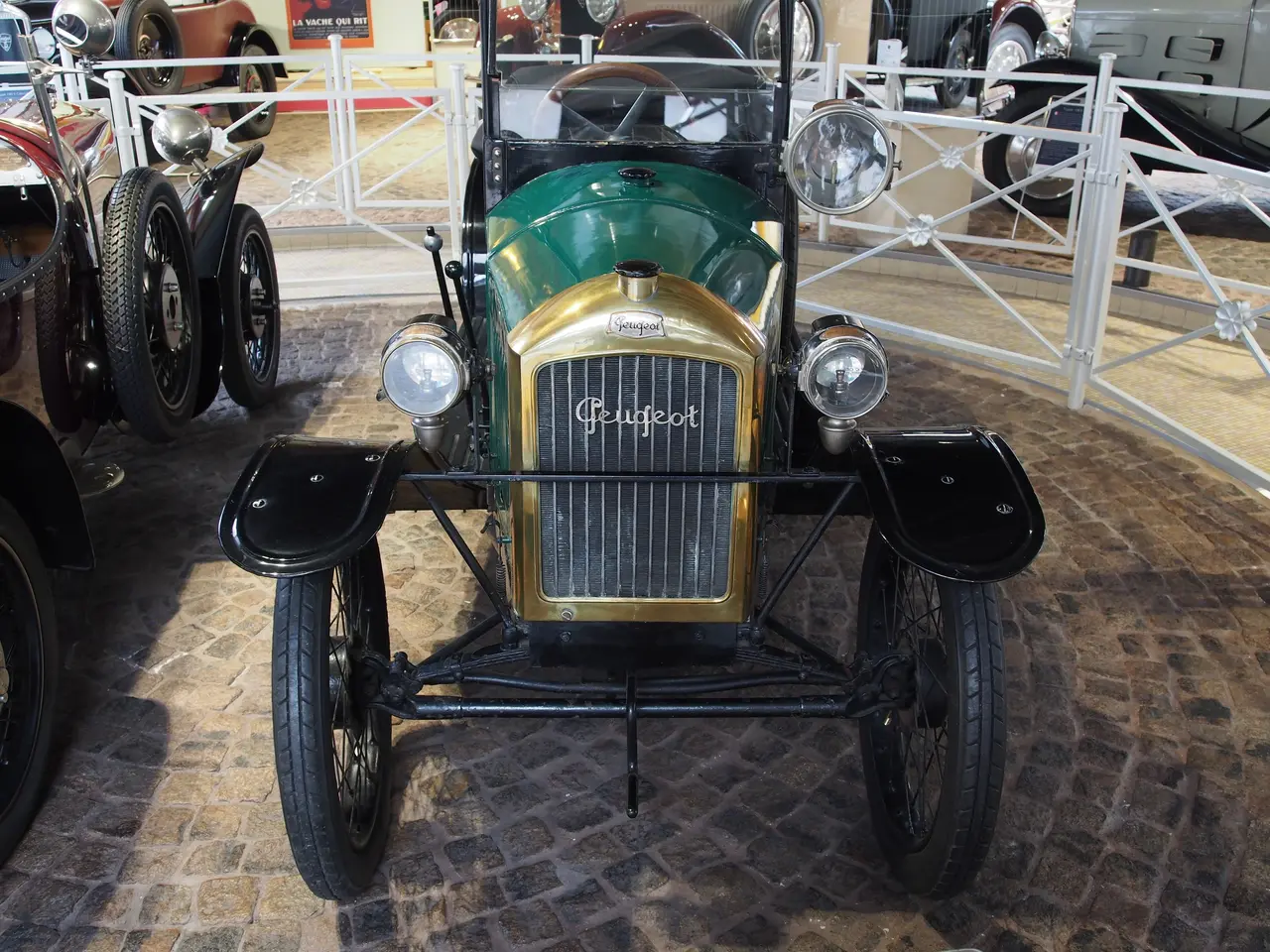 The image shows a vintage car on display in a museum, surrounded by other cars on the floor and a...
