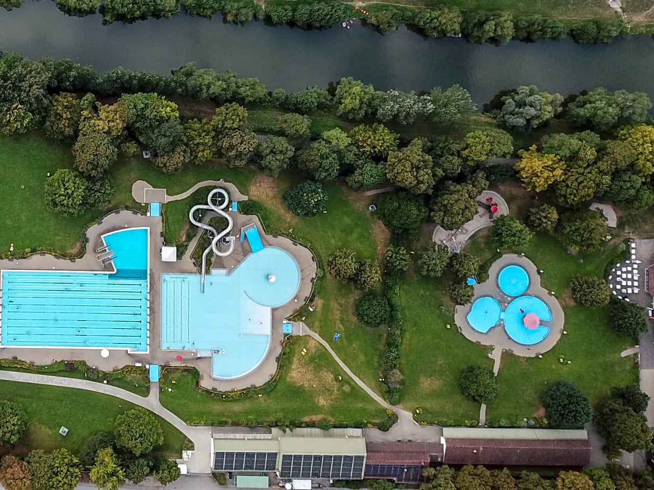 The image shows an aerial view of a park with two swimming pools surrounded by trees, grass, roads,...