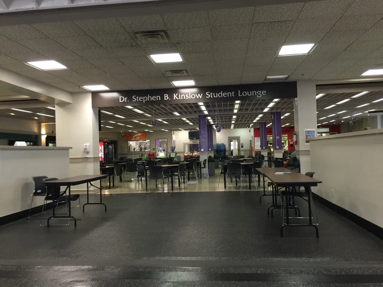 The image shows a large room filled with lots of tables and chairs, pillars, a name board, lights...