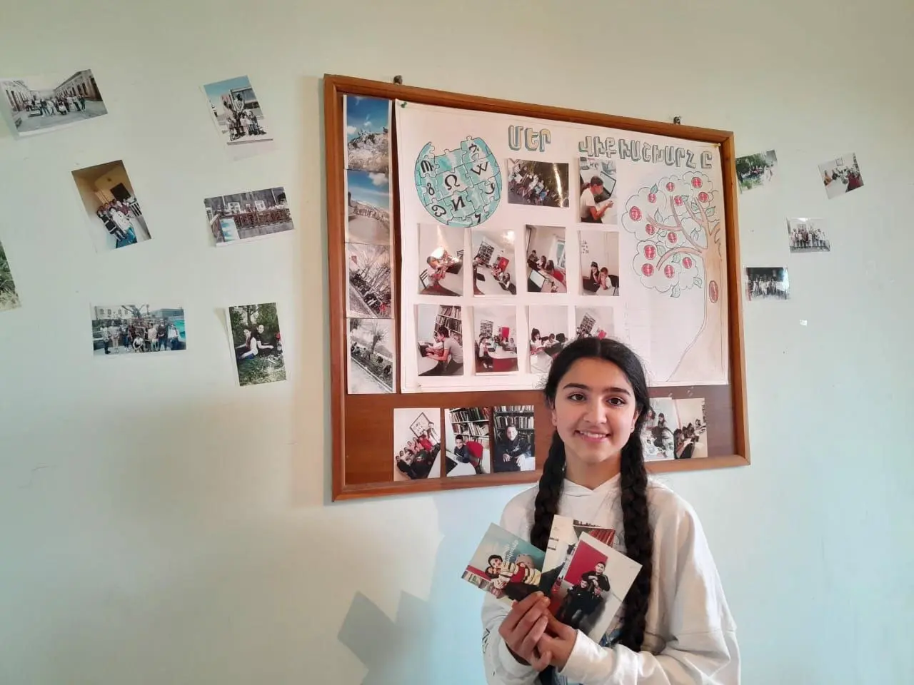 The image shows a young girl standing in front of a wall adorned with photos, holding a book in her...