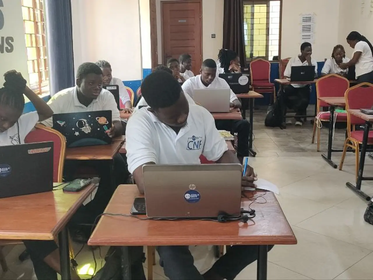 The image shows a group of people sitting at desks with laptops in a classroom. There are bags on...