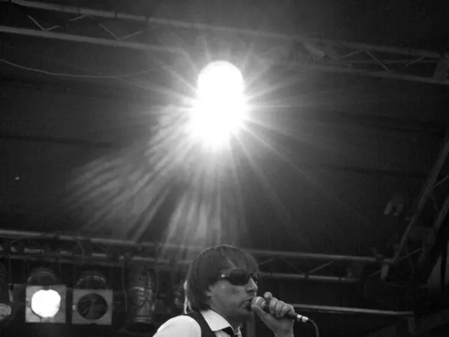 A man is singing with a mic in his hand. There is a man behind him holding a guitar. A light is...