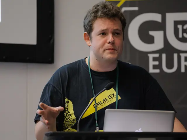 In the image there is a podium with mic and laptop. In front of that there is a man with black...