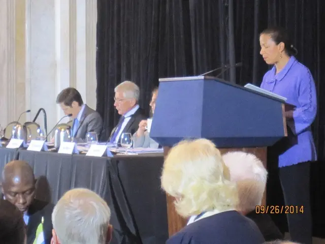 It is a conference, there is a woman standing in front of a table and speaking something, beside...