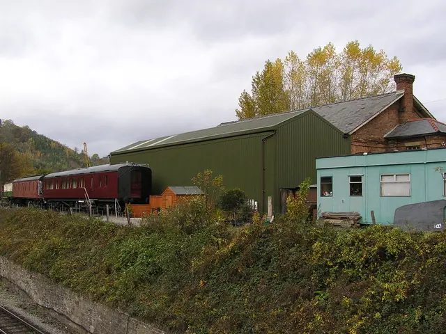 In this image in the front there are plants. In the center there is a train and there are sheds. In...