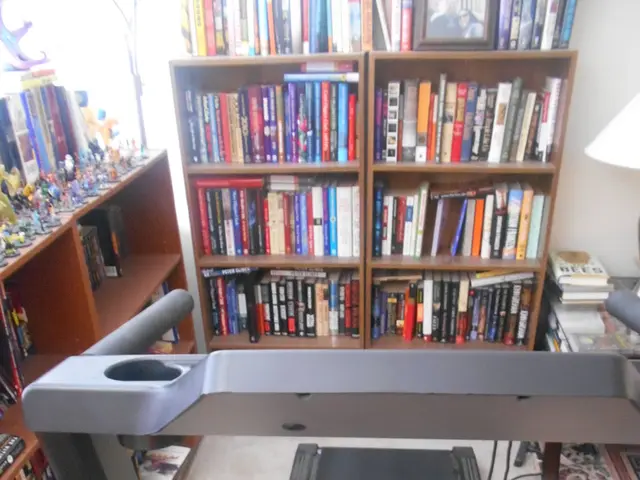 In the center of the image there is a treadmill. In the background of the image there are books...