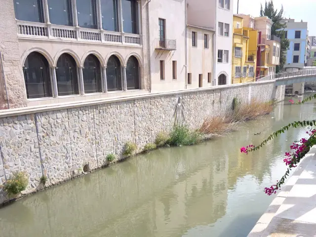 The image shows a small river running through a city next to tall buildings, with a wall on one...