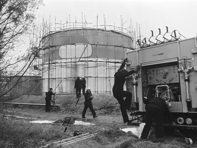 The image shows a group of men working on a truck in front of a large tank, surrounded by trees,...