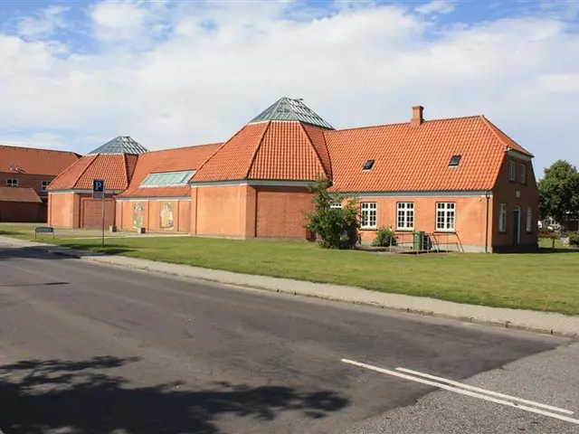 The image shows a large brick building with a red roof situated on the side of a road, surrounded...