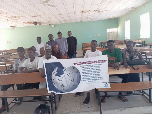 The image shows a group of people sitting at desks in a classroom, with a banner held up in front...