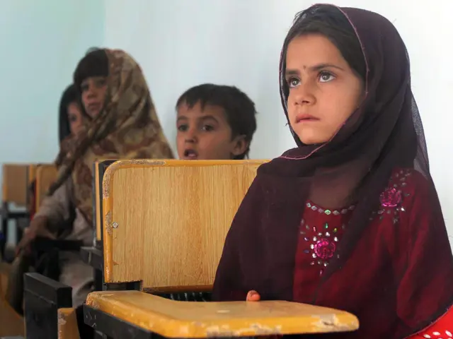 The image shows a group of children sitting at desks in a classroom, with a wall in the background....