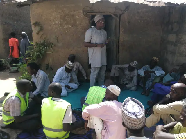 The image shows a group of people sitting on the ground in front of a building, with a man standing...