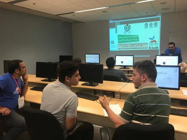 The image shows a group of people sitting at desks in front of computers, with monitors, keyboards,...