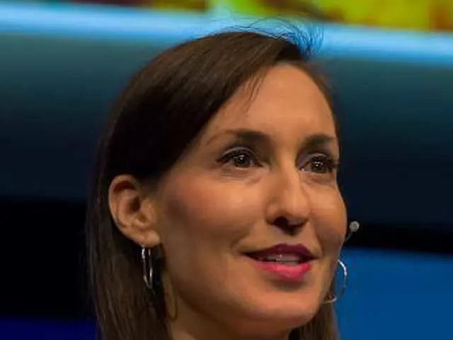The image shows a woman in a white jacket and black top sitting down in front of a blue and yellow...