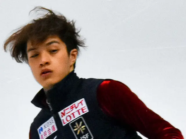 The image shows a young man in a black and red shirt skating on an ice rink, with a white...
