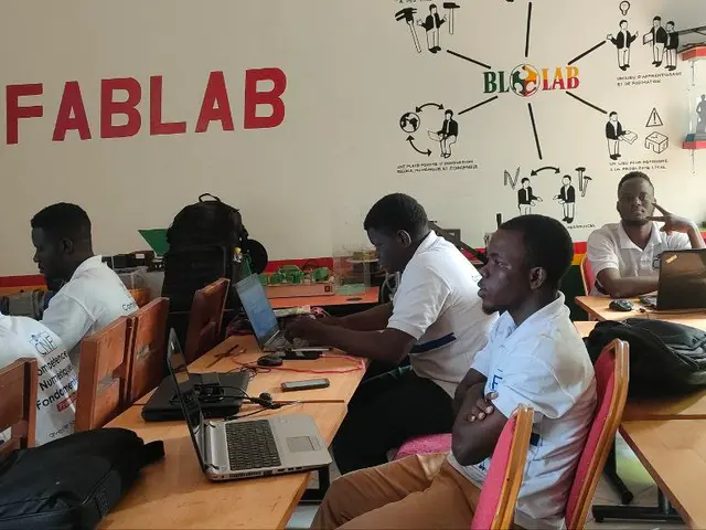 The image shows a group of people sitting at desks with laptops, bags, and other objects on the...