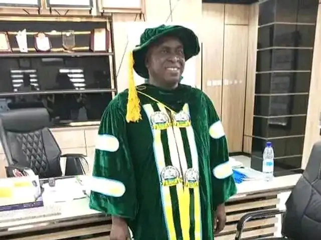 The image shows a man in a green graduation gown and hat standing in front of a desk, surrounded by...